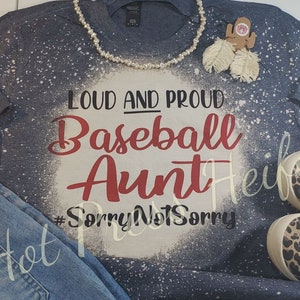 May include: A navy blue long-sleeved shirt with a distressed white design and the words "Loud and Proud Baseball Aunt" in red and white. The shirt is paired with blue jeans and leopard print sneakers. A pearl necklace and decorative earrings are also present.