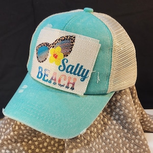 May include: A turquoise trucker hat with a distressed brim and a beige mesh back. The front patch features sunglasses, a yellow flower, and the words "Salty Beach" in a colorful font. A casual accessory for beach lovers.