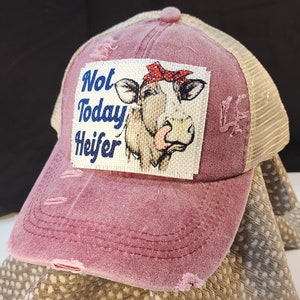 May include: A distressed maroon trucker hat with a beige mesh back. The front patch features a cow graphic with a red bandana and the text "Not Today Heifer" in blue. The hat has a curved brim and a pink button.