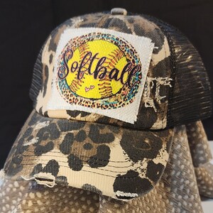 Könnte beinhalten: Eine Used-Trucker-Mütze mit Leopardenmuster und schwarzem Mesh-Rücken. Der vordere Aufnäher zeigt ein gelbes Softball-Design mit dem Wort "Softball" in Schreibschrift.