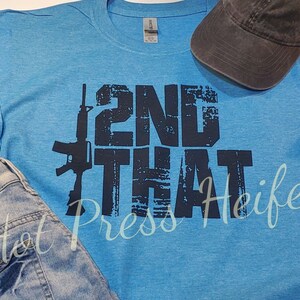 Puede incluir: Una camiseta azul claro con el texto "2ND THAT" y un gráfico de rifle en azul oscuro. Una gorra de béisbol gris oscuro y unos vaqueros azules también están en la imagen. Las palabras "Hot Press Heifer" están escritas en cursiva.
