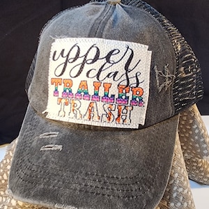 May include: Distressed grey trucker hat with a black mesh back. The hat features a patch with the words "upper class trailer trash" in colourful lettering. The hat has a pre-curved peak and a button on top.
