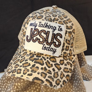 May include: A leopard print trucker hat with a mesh back. The hat has a patch with the text "only talking to JESUS today" in black and orange lettering. The hat is in shades of tan and brown.