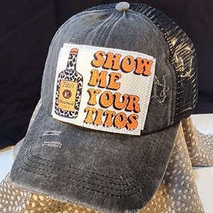 May include: Distressed grey trucker hat with a black mesh back. The front patch features a Tito's Vodka bottle and the phrase "SHOW ME YOUR TITOS" in orange letters. The hat has a pre-worn appearance.