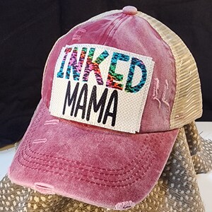 May include: A distressed, dusty rose baseball cap with a mesh back. The front features a white patch with the words "INKED MAMA" in black and colorful lettering. The cap has a curved brim and a button on top.