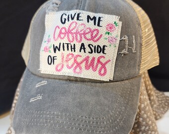 May include: Gray distressed trucker hat with a mesh back. The front patch reads "GIVE ME coffee WITH A SIDE OF Jesus" in pink script and black block letters, with floral accents. The hat has a curved brim.