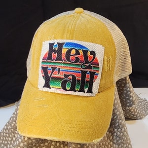 May include: Mustard yellow baseball cap with a distressed look. The front features a patch with the text "Hey Y'all" in a colorful, retro design. The cap has a mesh back and a curved brim.