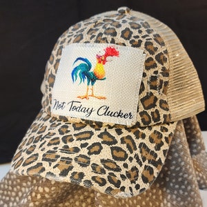 Può includere: Cappellino da camionista con stampa leopardata e retro in rete. La toppa anteriore presenta un'illustrazione di gallo colorato con la scritta "Not Today Clucker". Il cappellino ha una visiera curva e una chiusura a scatto.