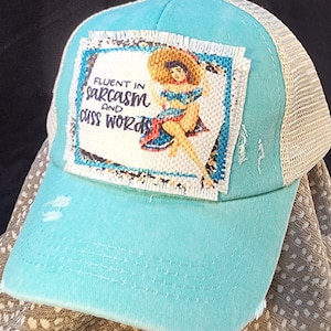 Peut inclure: Casquette de camionneur turquoise avec un dos en filet blanc. L'avant présente un écusson avec le texte "Fluent in Sarcasm and Cuss Words" et une illustration de pin-up. La casquette a un aspect vieilli.