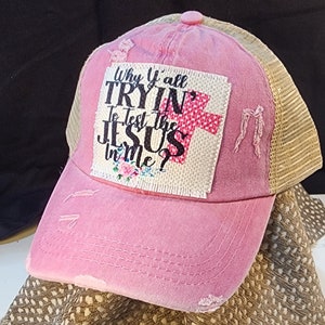 Op de afbeelding: Roze distressed baseballcap met een mesh achterkant. De voorkant heeft de tekst "Why Y'all Tryin' To Test The Jesus in Me?" met een kruis en bloemenaccenten. De cap heeft een gebogen klep en een knoop bovenop.