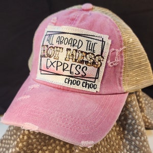 May include: Pink distressed trucker hat with a tan mesh back. The front patch reads "ALL ABOARD THE HOT MESS EXPRESS choo choo" in black and gold lettering. The hat has a curved brim and a button on top.