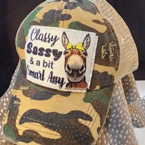 May include: Camouflage trucker hat with a mesh back and a patch featuring a donkey with a yellow bow and the text "Classy, Sassy & a bit Smart Assy". The hat has a curved brim and a button on top.