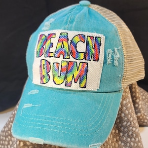 Pode incluir: Boné de beisebol turquesa com parte traseira em malha bege e detalhes desgastados. A frente apresenta um patch com as palavras "BEACH BUM" em um design tie-dye arco-íris. O boné tem uma viseira curva e um botão no topo.