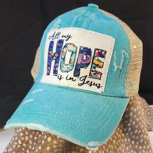 Puede incluir: Gorra de béisbol turquesa desgastada con parte trasera de malla. La parte delantera presenta un parche blanco con las palabras "All my HOPE is in Jesus" en un diseño floral. La gorra tiene una visera precurvada.