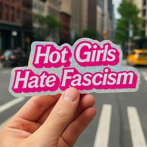 May include: A holographic sticker with the text "Hot Girls Hate Fascism" in bright pink, outlined in white. The sticker is held up in front of a city street background.