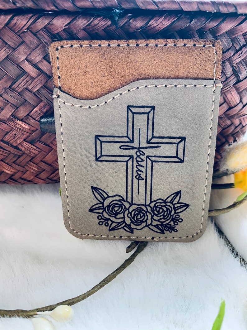 May include: A tan leather cardholder with a cross design. The cross is outlined in black, with the word "Jesus" written vertically through the center. Floral accents are at the base of the cross. The top pocket is a lighter brown.