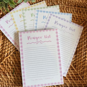 May include: A stack of pastel-colored prayer list notepads with a gingham border. Each notepad features the words "Prayer List" in a script font, with lines for writing. The notepads are arranged on a woven wicker surface.