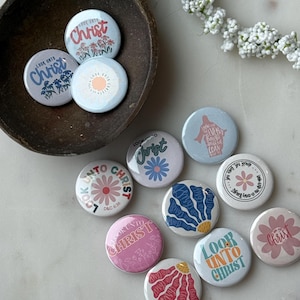 May include: A collection of round, colorful buttons with the phrase "Look Unto Christ" and floral designs. The buttons are in various colors, including pink, blue, and white, and are arranged on a marble surface.