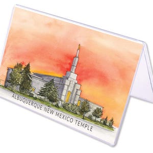 May include: A clear plastic display stand featuring a watercolor painting of the Albuquerque New Mexico Temple. The artwork depicts the temple with a vibrant orange and yellow sunset in the background. The text "ALBUQUERQUE NEW MEXICO TEMPLE" is printed at the bottom.