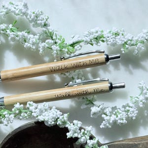 May include: Two bamboo pens with silver accents and the engraved text "Walk With Me." The pens are set against a backdrop of white floral accents and a dark brown, textured surface. The pens are a light brown color.