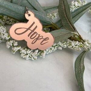 May include: A rose gold colored metal keychain with the word "Hope" written in cursive on it. The keychain is hanging from a silver chain and is surrounded by white flowers and green leaves.