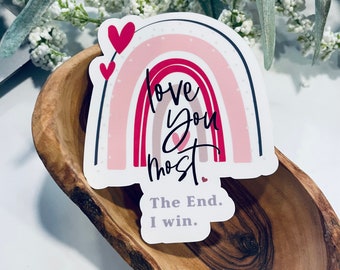 May include: A pink and white rainbow sticker with the text "love you most" in black script. The sticker has two small red hearts on either side of the rainbow. The sticker is on a wooden surface. The text "The End. I win." is below the rainbow.