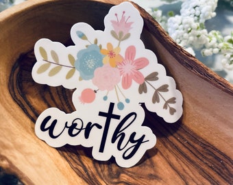 May include: A sticker with a floral design in pastel colors, including pink, blue, and yellow, with the word "worthy" in black script. The sticker is set in a wooden bowl with a floral background.