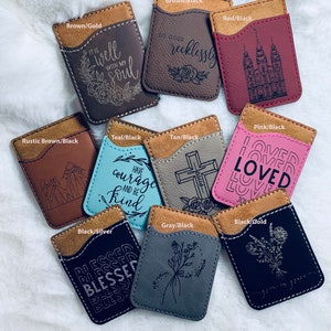 May include: A collection of leather phone wallets in various colors, including brown, teal, pink, and black. Each wallet features a contrasting suede-like top and engraved designs with inspirational quotes and floral motifs. The wallets are designed to hold cards.