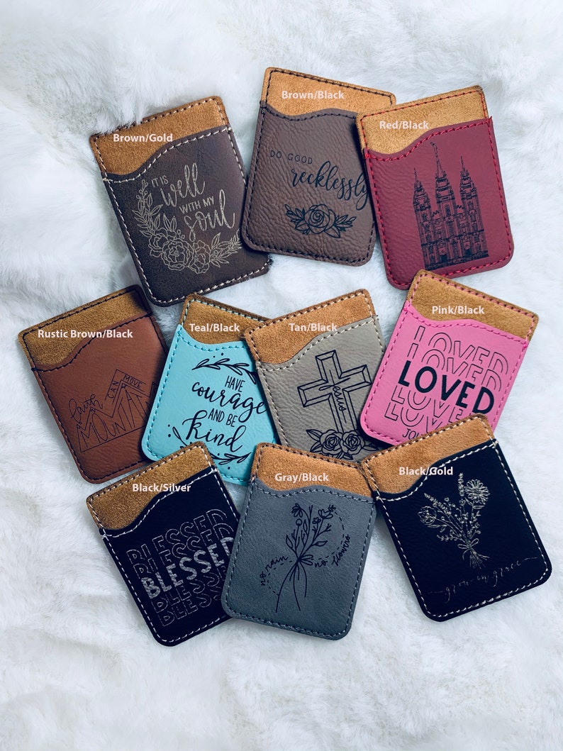 May include: A collection of leather phone wallets in various colors, including brown, teal, pink, and black. Each wallet features a contrasting suede-like top and engraved designs with inspirational quotes and floral motifs. The wallets are designed to hold cards.