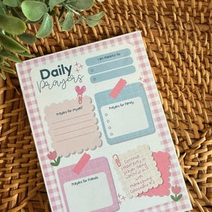 May include: A Daily Prayers notepad with a pink gingham pattern. The notepad includes sections for prayers for self, family, and friends, with the text "I am thankful for" and a Bible verse. The design features pink and blue accents.