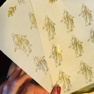 May include: Two sheets of white paper with gold foil stickers of a woman in a flowing robe. The stickers are arranged in rows on the paper. The paper has the text "lily ann designs" printed in gold foil at the top.