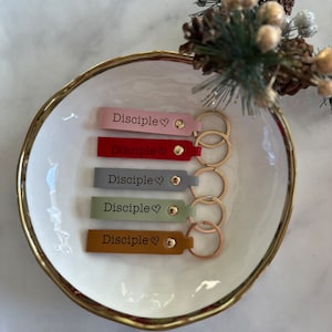 May include: Five leather keychains with gold rings. Each keychain has the word "Disciple" and a heart symbol engraved on it. The keychains are in different colors: pink, red, gray, green, and brown.