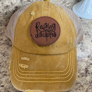 May include: A mustard yellow distressed baseball cap with a brown leather patch that says "Raising Tiny Disciples".
