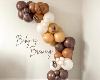 Puede incluir: Una guirnalda de globos en tonos marrón, beige y blanco, dispuesta en forma de arco contra una pared blanca. El texto "Baby is Brewing" está escrito en cursiva. Los globos tienen acabados mate y brillantes.