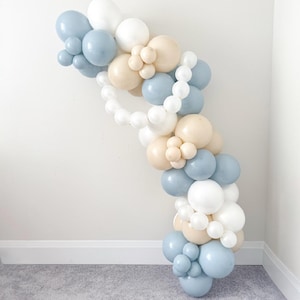 May include: A balloon arch in a soft color palette of white, light blue, and beige. The balloons are arranged in a cascading, curved shape, perfect for a party or event decoration. The balloons are of varying sizes.