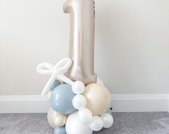 Op de afbeelding: Een ballonnenarrangement voor een feest. Een grote, metallic ballon in de vorm van het cijfer één in een neutrale tint staat centraal. De basis bestaat uit witte, lichtblauwe en crèmekleurige ballonnen, met een witte strik. Perfect voor een eerste verjaardag.