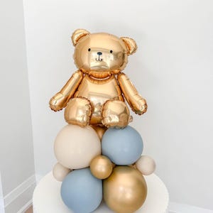 Peut inclure: Un ballon en forme d'ours doré est posé sur un ensemble de ballons crème, bleus et dorés. L'ours a un visage souriant et est placé sur une table ronde blanche. Idéal pour une baby shower ou un anniversaire.