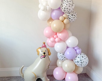 May include: A festive balloon arch in pastel colors, including pink, white, and lavender, with paw print accents. A large, smiling dog-shaped balloon with a party hat sits nearby, creating a celebratory atmosphere.