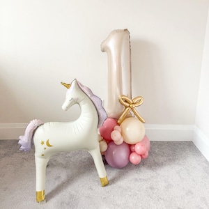 May include: A white unicorn balloon with gold accents and a pink and purple mane stands next to a balloon arrangement. The arrangement includes a large number one balloon, a gold bow, and pink and cream-colored balloons. A festive decoration.