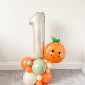 May include: A festive balloon arrangement for a first birthday. It features a large silver number one balloon, an orange smiling face balloon, and a cluster of orange, green, and cream-colored balloons at the base. The balloons are arranged on a gray carpet.