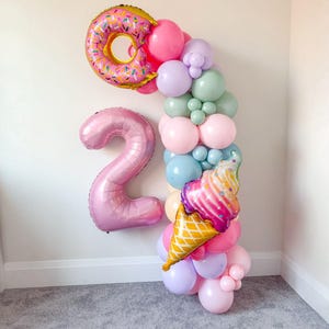 May include: A vibrant balloon arch featuring a pink donut, the number 2, and an ice cream cone. The arch is composed of pink, purple, blue, and green balloons, ideal for a birthday party. The ice cream cone balloon has a waffle cone base.