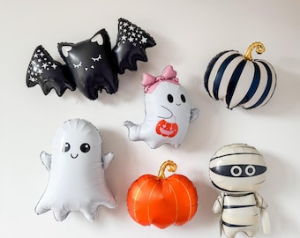 May include: A collection of Halloween-themed foil balloons including a black bat with white stars, a white ghost with a pink bow holding an orange pumpkin, a blue and white striped pumpkin, a white ghost, an orange pumpkin, and a white mummy.