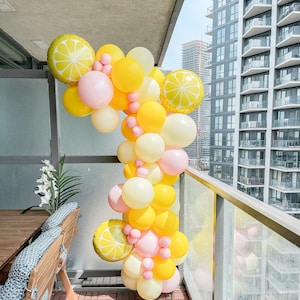 Könnte beinhalten: Ein Ballonbogen aus gelben, rosa und weißen Ballons. Die Ballons sind in Form von Zitronen geformt und in einem kaskadierenden Muster angeordnet. Der Bogen befindet sich auf einem Balkon mit Blick auf eine Skyline.