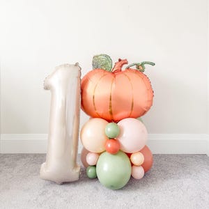 May include: A celebratory balloon display. A large peach-coloured pumpkin balloon with green detailing sits atop a cluster of pastel balloons. A large, silver number "1" balloon is positioned beside the pumpkin, ideal for a first birthday.