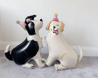 May include: Two dog-shaped balloons. One is black and white, wearing a pink and white striped party hat. The other is cream-colored with a pink polka dot party hat. Both balloons are sitting on a gray carpet.
