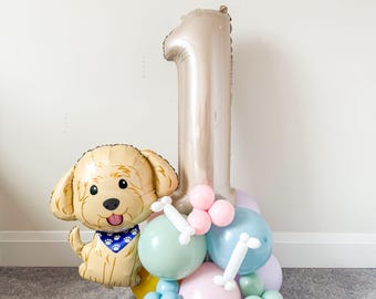May include: A festive balloon arrangement featuring a large number one balloon in a metallic gold color. A dog-shaped balloon with a blue bandana sits next to the number. The base includes pastel-colored balloons in pink, blue, and green.