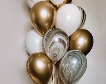 May include: A bouquet of balloons in white, gold, and marble patterns. The balloons are tied together with gold ribbons, creating a festive arrangement. The balloons are a mix of solid colors and swirled designs.