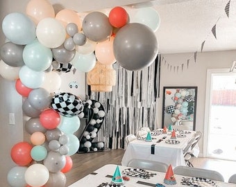 May include: A birthday party table setting with a checkered tablecloth, black and white checkered plates, and turquoise party hats. A balloon arch made of gray, white, peach, and turquoise balloons is in the background.