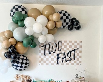 May include: A birthday party table setting with a checkered banner that says "Two Fast". The table is decorated with black, white, and green balloons, and there are various snacks and desserts on the table, including a two-tiered cake with a race car theme.