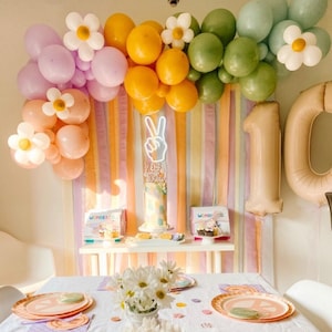 May include: A birthday party table setting with a white tablecloth, colourful plates featuring peace signs, and white daisies in mason jars. The backdrop features a rainbow of streamers and balloons with daisies. The cake is decorated with a peace sign and the words "Let's Get Funky 10 Years".